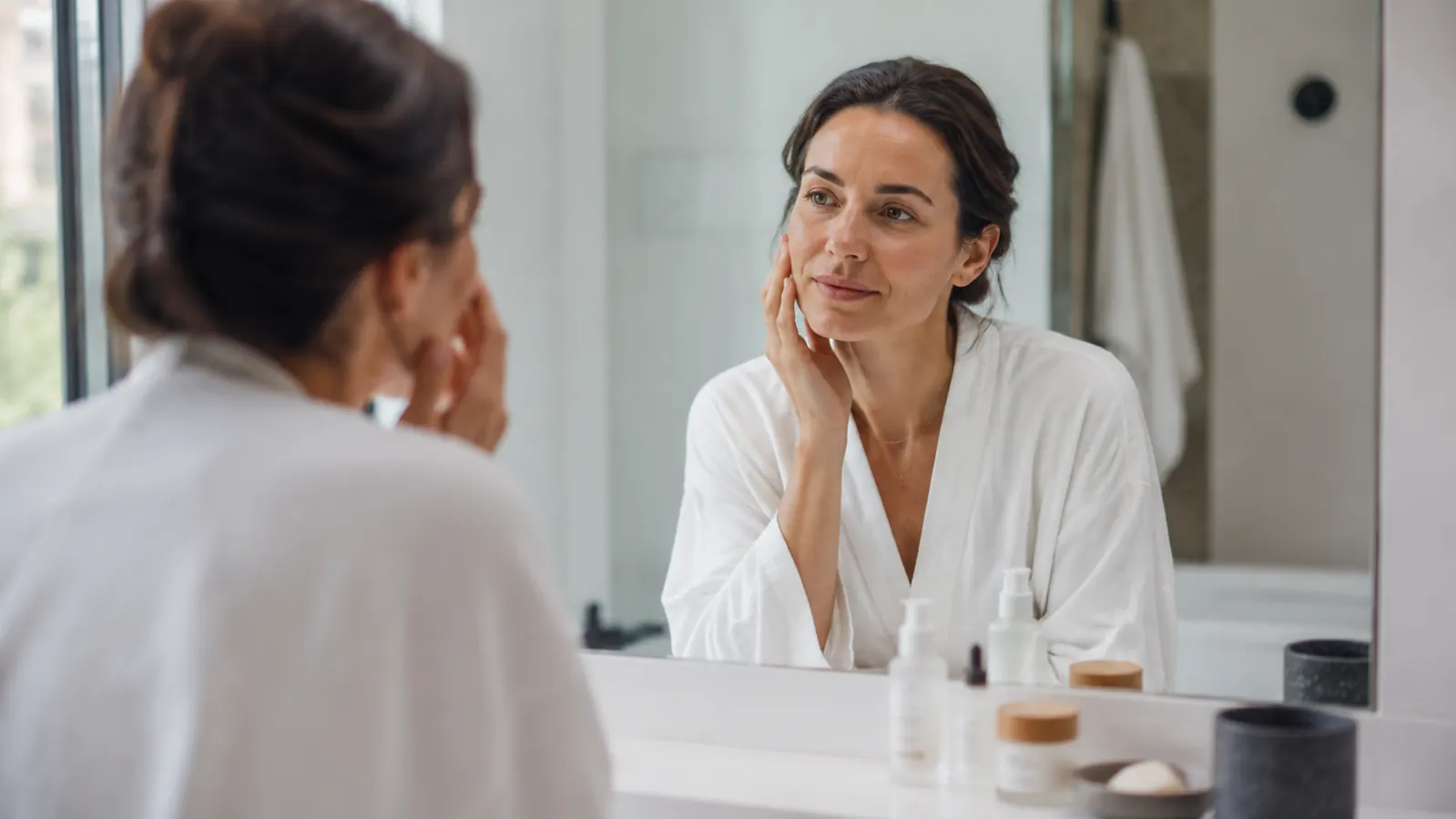 Woman in her late 30s sitting at a bathroom vanity in morning light, applying skincare, looking calm and rested