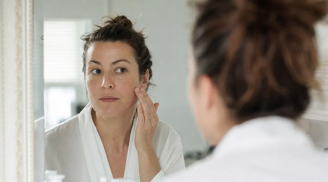 Woman touching her cheek and examining skin texture in a well-lit bathroom mirror